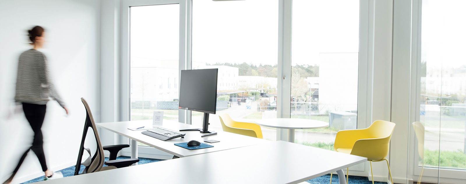 Ein modernes Büro mit einem Schreibtisch, einem Computerbildschirm und einem gelben Stuhl. Eine Person geht im Hintergrund vorbei.