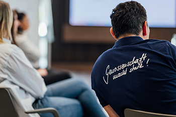 Galeriebild VEMA_2357.jpg Eine Person von hinten, die ein T-Shirt mit dem Schriftzug „Gewissenschaft macht stark!“ trägt, sitzt vor einer Gruppe von Menschen in einem Konferenzraum.