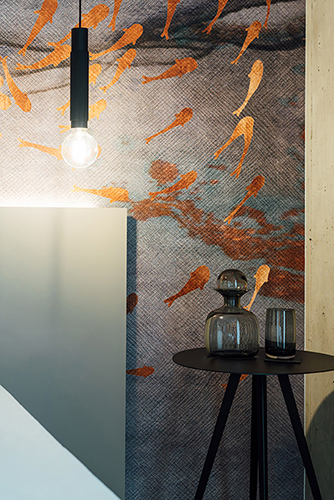 Galeriebild VEMA_2365.jpg Ein minimalistisch gestalteter Raum mit einem von Fischen dekorierten Wandmuster, einer hängenden Lampe und einem Tisch mit Glasgefäßen.
