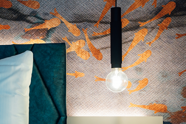Galeriebild VEMA_2368.jpg Ein Nachttisch mit einer Glühbirne und einem weißen Kopfkissen vor einer Wand mit Fischmotiven.