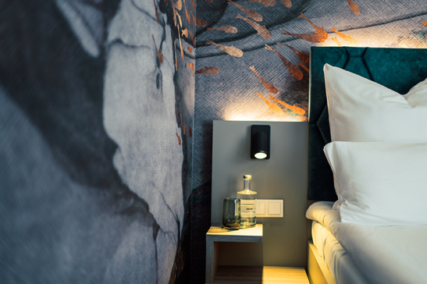 Galeriebild VEMA_2548.jpg Ein modern eingerichtetes Schlafzimmer mit einer dekorativen Wand, einem Nachttisch, einer Lampe und Wasserflaschen.