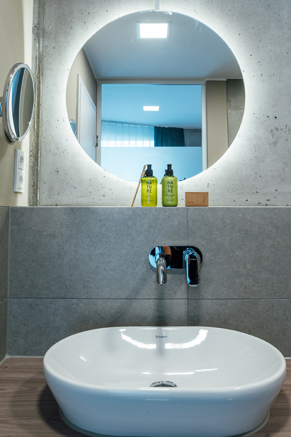 Galeriebild VEMA_2767.jpg Ein modernes Badezimmer mit einem runden Spiegel, einem Waschbecken und zwei grünen Shampoo-Flaschen.