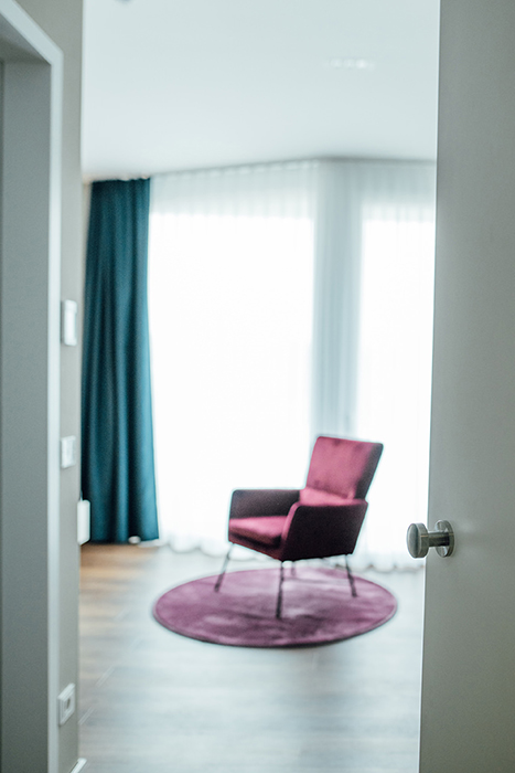 Galeriebild VEMA_2812.jpg Ein offenes Zimmer mit einem lila Stuhl auf einem runden Teppich und Vorhängen, die Licht hereinlassen.