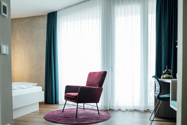 Galeriebild VEMA_2823.jpg Ein stilvolles Zimmer mit einem lila Sessel, einem runden Teppich und großen Fenstern mit Vorhängen.