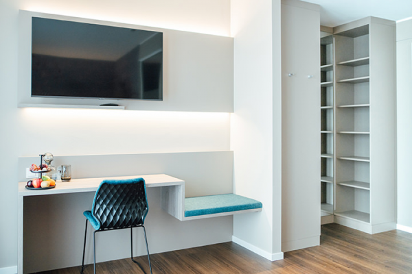 Galeriebild VEMA_2912.jpg Moderne Hotelzimmer mit einem Fernseher an der Wand, einem Schreibtisch, einem Stuhl und einem offenen Regal.