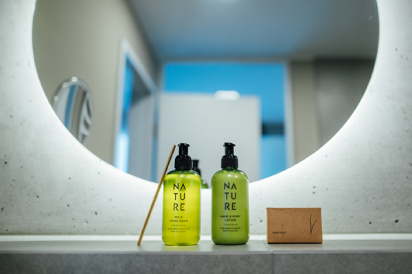 Galeriebild VEMA_2966.jpg Zwei grüne Flaschen mit Seife und Lotion, eine Holzstange und eine kleine braune Box auf einem Waschbecken vor einem Spiegel.