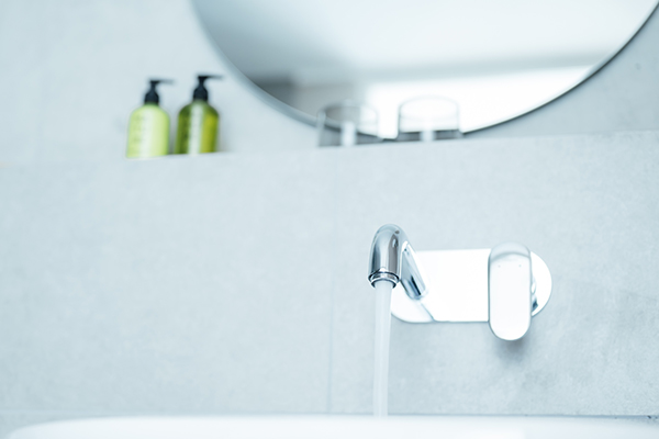 Galeriebild VEMA_3519.jpg Ein Wasserhahn mit fließendem Wasser in einem modernen Badezimmer, im Hintergrund sind Pflegeprodukte und ein Spiegel sichtbar.