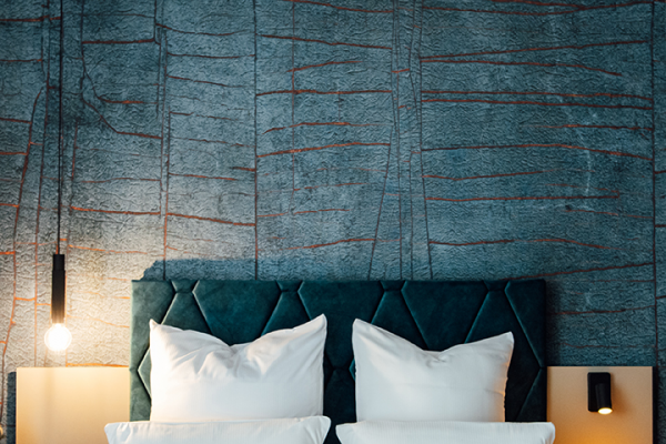 Galeriebild VEMA_3621.jpg Ein Kopfteil eines Bettes mit zwei Kissen vor einer strukturierten Wand in Blau- und Rottönen. Zwei bedside lamps sind sichtbar.