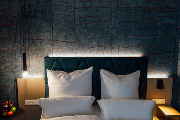 Galeriebild VEMA_3677.jpg Ein modernes Hotelzimmer mit einem gepolsterten Bettkopfteil, weißen Kissen, einer laminierten Wand und einer Nachttischlampe.