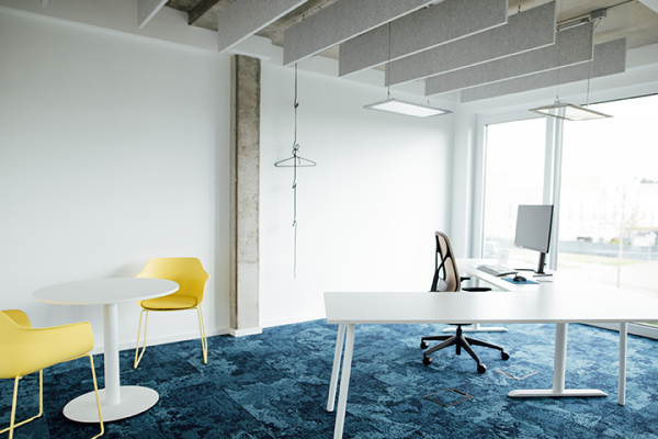 Galeriebild VEMA_3817.jpg Ein modernes Büro mit einem weißen Tisch, einem Bürostuhl, zwei gelben Stühlen und einem Computer am Tisch. Der Boden ist blau und es gibt große Fenster.