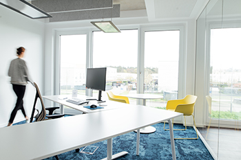 Galeriebild VEMA_4029.jpg Ein modernes Büro mit einem Schreibtisch, einem Computer, Stühlen und großen Fenstern. Eine Person läuft im Hintergrund.