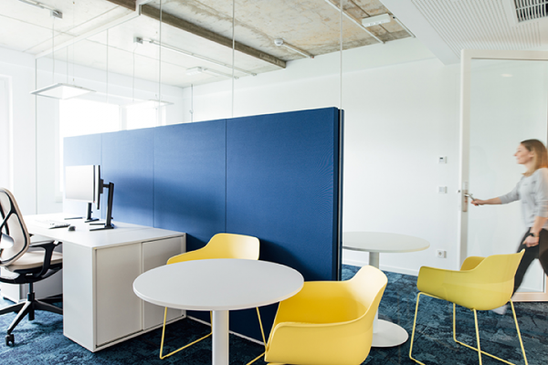 Galeriebild VEMA_4182.jpg Ein modernes Büro mit einem Schreibtisch, zwei gelben Stühlen, einem weißen Tisch und einer blauen Trennwand. Eine Person betritt den Raum.
