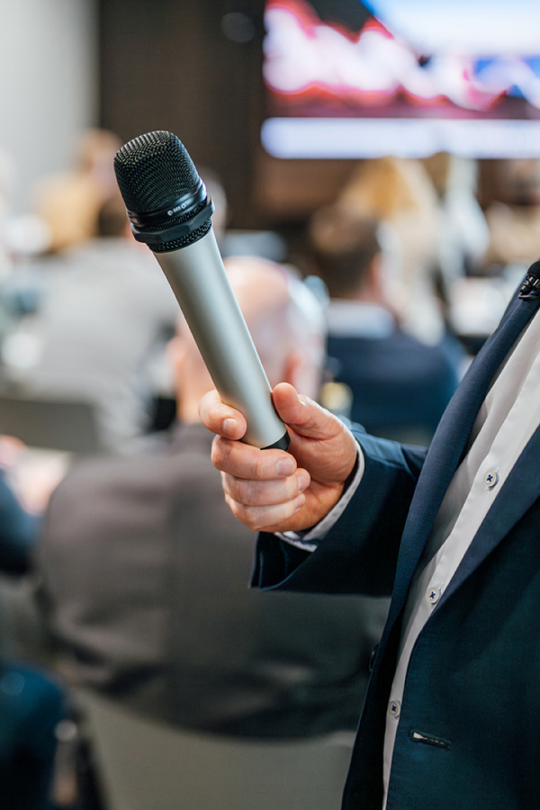 Galeriebild VEMA_4576.jpg Ein Mikrofon wird von einer Hand gehalten, Hintergrund mit unscharfen Personen und Präsentation.