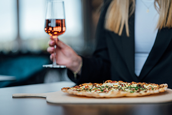 Galeriebild VEMA_5377A.jpg Eine Person mit einem Glas Roséwein in der Hand sitzt an einem Tisch mit einer Pizza.