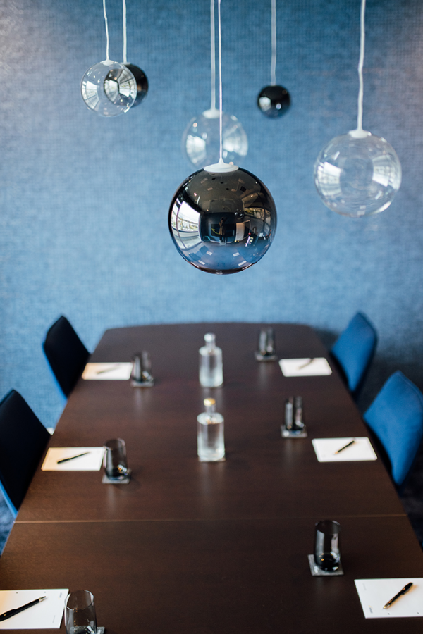 Galeriebild VEMA_6855.jpg Ein Konferenzraum mit einem langen Tisch. Über dem Tisch hängen mehrere runde Lampen. Auf dem Tisch stehen Gläser, Wasserkrüge und Schreibunterlagen. Die Wände sind blau.