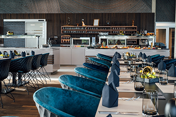 Galeriebild VEMA_8384.jpg Ein modernes Restaurant mit eleganten blauen Stühlen, einem langen Tisch, dekorativen Elementen und einem Buffetbereich im Hintergrund.