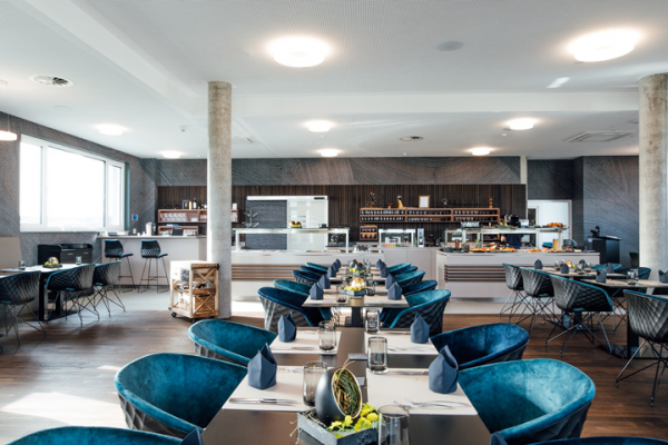 Galeriebild VEMA_8618.jpg Ein modernes Restaurant mit eleganten blauen Stühlen, gedeckten Tischen und einer gemütlichen Atmosphäre. Im Hintergrund befindet sich eine Theke mit Essensangeboten.