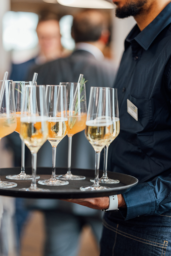 Galeriebild VEMA_8645.jpg Eine Person trägt ein Tablett mit Gläsern, die verschiedene Getränke enthalten, darunter Sekt und Cocktails mit Rosmarin.