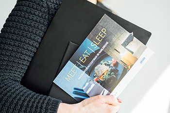 Galeriebild VEMA_9825.jpg Ein Mensch hält eine schwarze Mappe und eine Broschüre mit dem Titel „MEET EAT SLEEP“.
